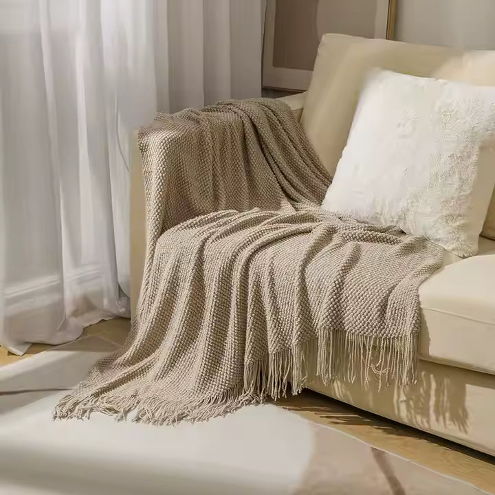 Beige blanket draped over a beige sofa with white cushions in a living room setting.