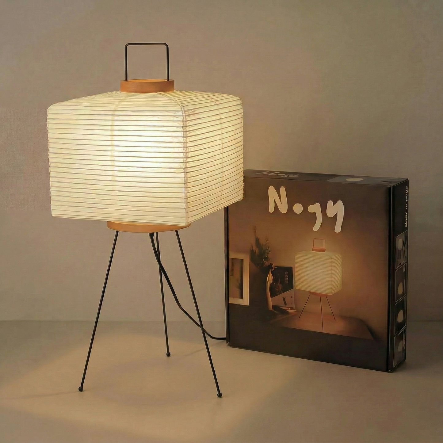 Illuminated square Japanese paper floor lamp on a black stand next to its Nogy-branded packaging box in a minimalist indoor setting.