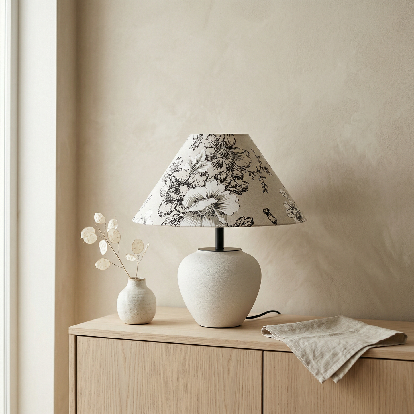 Minimalist japandi ceramic table lamp with white rounded base and a flower linen lampshade on a light wood console