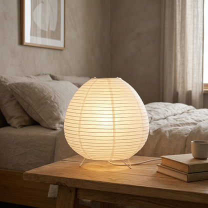 Japandi paper table lamp with a spherical rice paper shade emitting a warm glow on a wooden nightstand in a cozy minimalist bedroom.