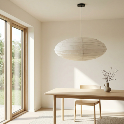 White paper lantern pendant lamp hanging over a wooden dining table in a bright, minimalist room with large glass windows and natural greenery.